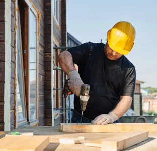 Construction worker in a yellow hard hat drilling into wooden boards outdoors near a building.