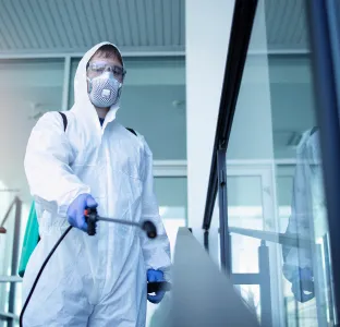 Person in white protective suit and mask disinfecting an indoor glass railing with spraying equipment.