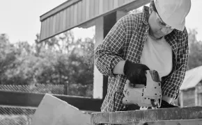 Construction worker wearing helmet and gloves using a power saw on a wooden board outdoors.