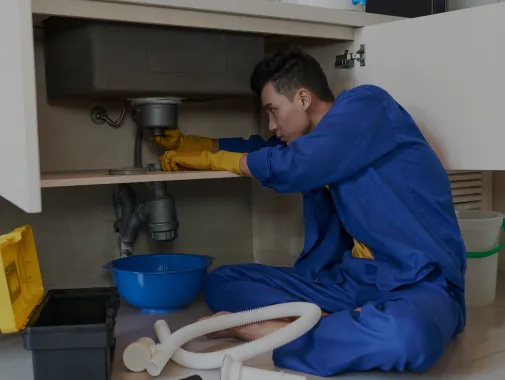 Plumber in blue coveralls and yellow gloves fixing a kitchen sink pipe with tools and bucket nearby.