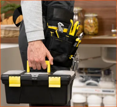 Person carrying a black and yellow toolbox with tools in a belt pouch against a blurred indoor background.