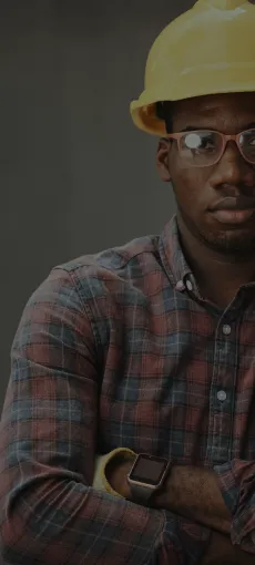 Man wearing a yellow construction helmet, glasses, a plaid shirt, and a smartwatch with arms crossed against a gray background.