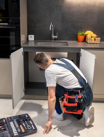 Plumber in overalls kneeling and inspecting under a kitchen sink with an open toolbox nearby.