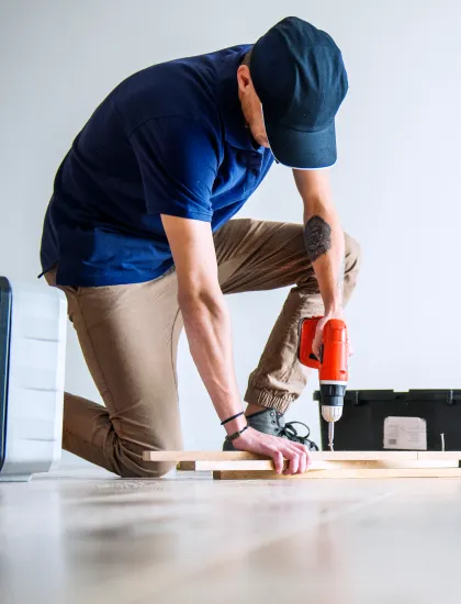 Person wearing a cap and blue shirt kneeling and using an electric drill on a wooden plank indoors.