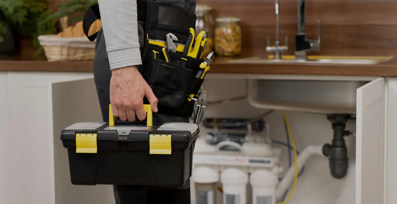 Person holding a black and yellow toolbox standing in front of an open kitchen cabinet with water filtration system and pipes visible underneath the sink.