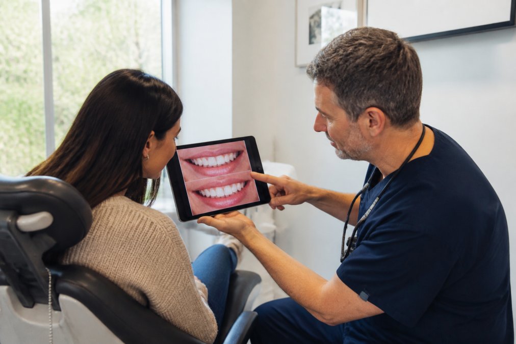 Cosmetic dentist showing a patient their AI-generated smile simulation on a tablet during a chairside consultation, with the patient leaning in to examine the digital preview while the dentist points to specific features
