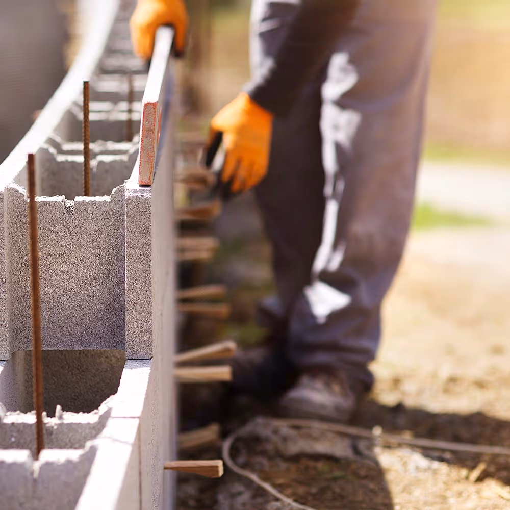 Construction worker wearing orange gloves using a level tool to align concrete blocks for a wall.