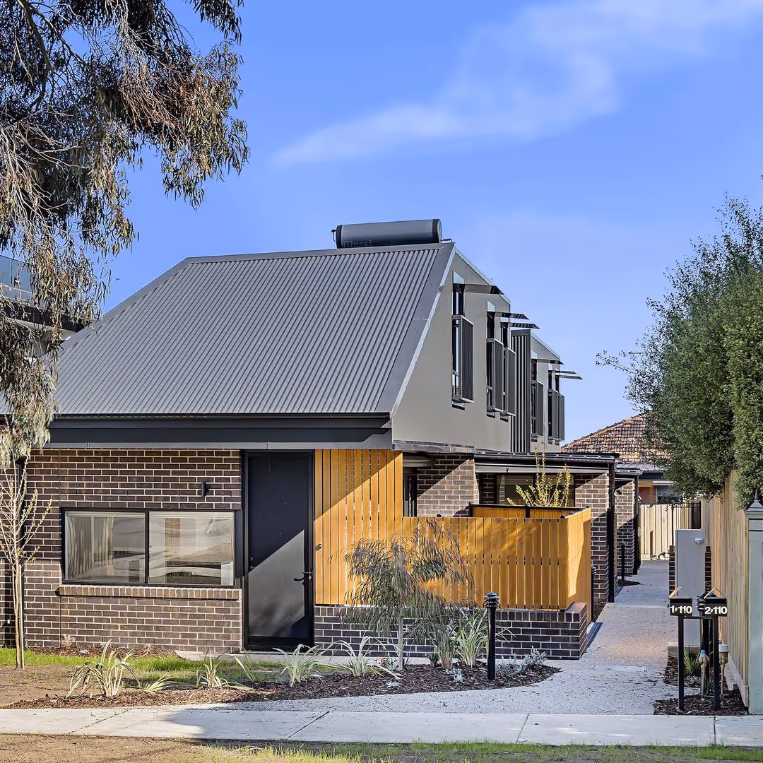 Modern two-story townhouse with dark brick exterior, black metal roof, and wooden fence under clear blue sky.