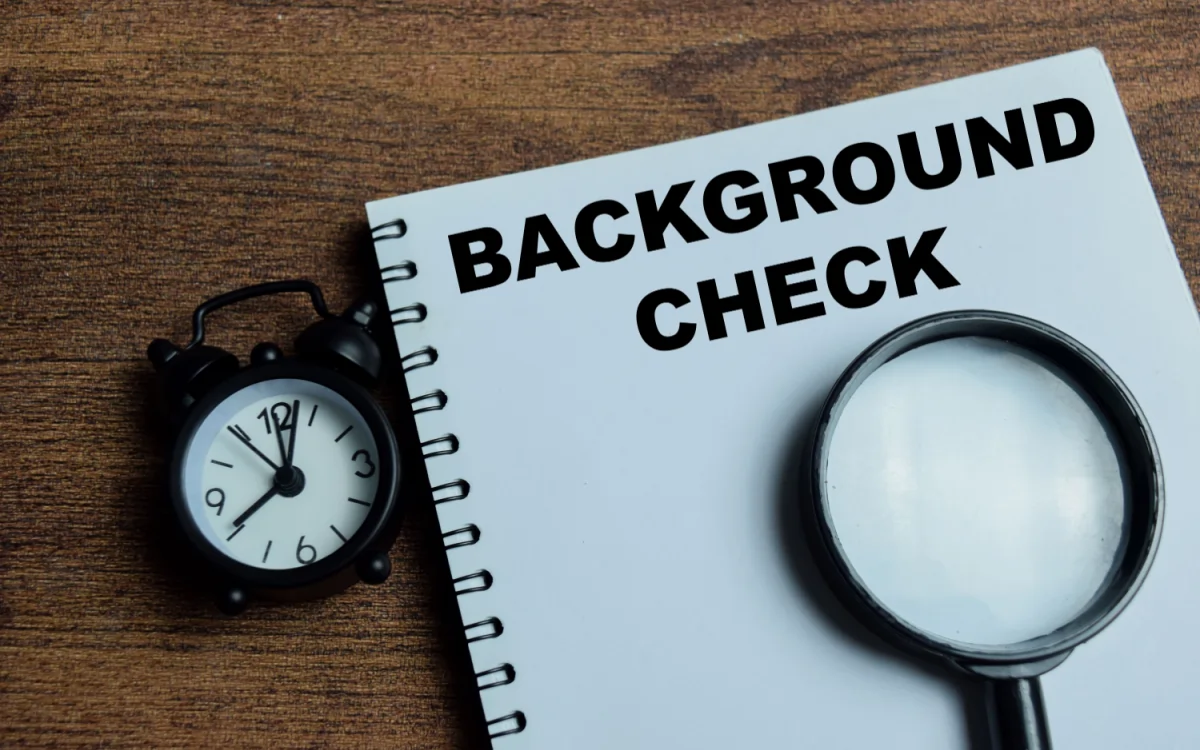 Notebook labeled “Background Check” with magnifying glass and clock, representing pre-employment screening, verification, and hiring risk assessment for employers.