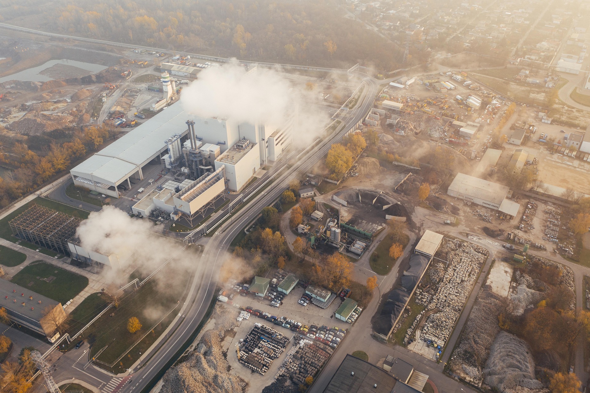 Aerial view of an industrial facility with large buildings, smokestacks emitting white smoke, and surrounding storage areas and roads.