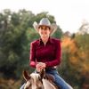 Smiling woman in a cowboy hat riding a horse with green trees and autumn foliage in the background.