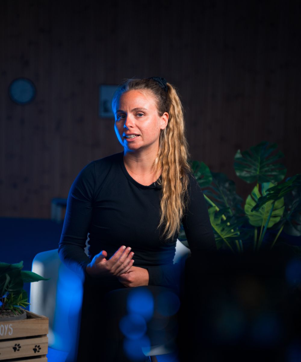 Woman with long blonde hair in a ponytail, wearing a black long-sleeve shirt, sitting and gesturing with her hand indoors.