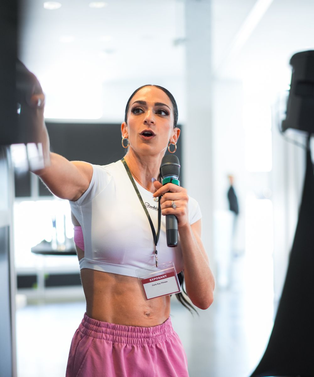 Fit woman in a white crop top and pink pants speaking into a microphone at a fitness event, wearing an exposant badge.