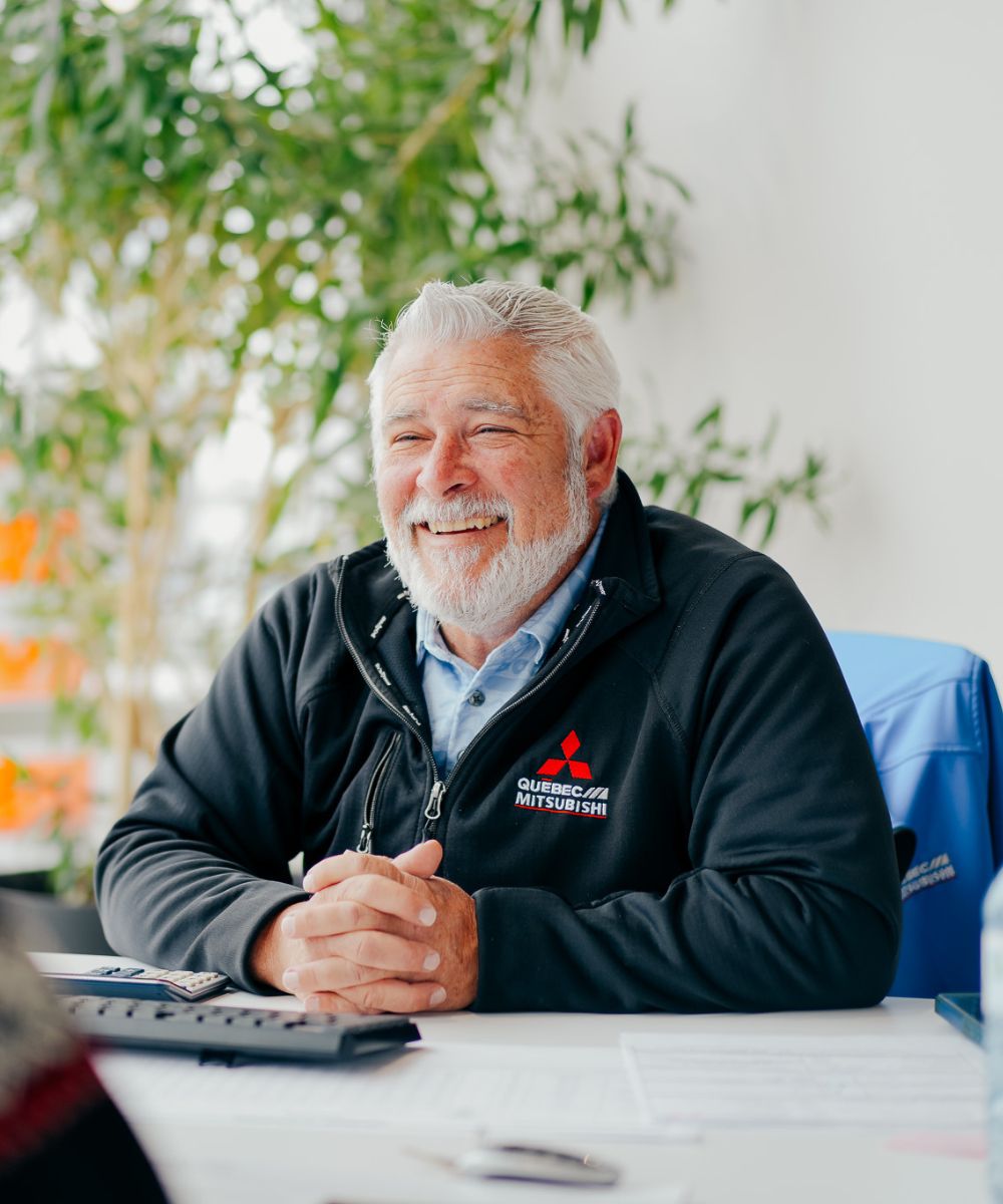 Smiling elderly man with white hair and beard wearing a Quebec Mitsubishi jacket seated at a desk.