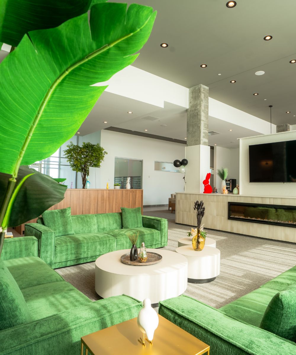 Modern living room with green velvet sofas, white circular coffee tables, large tropical leaves, and a wall-mounted TV above a built-in fireplace.