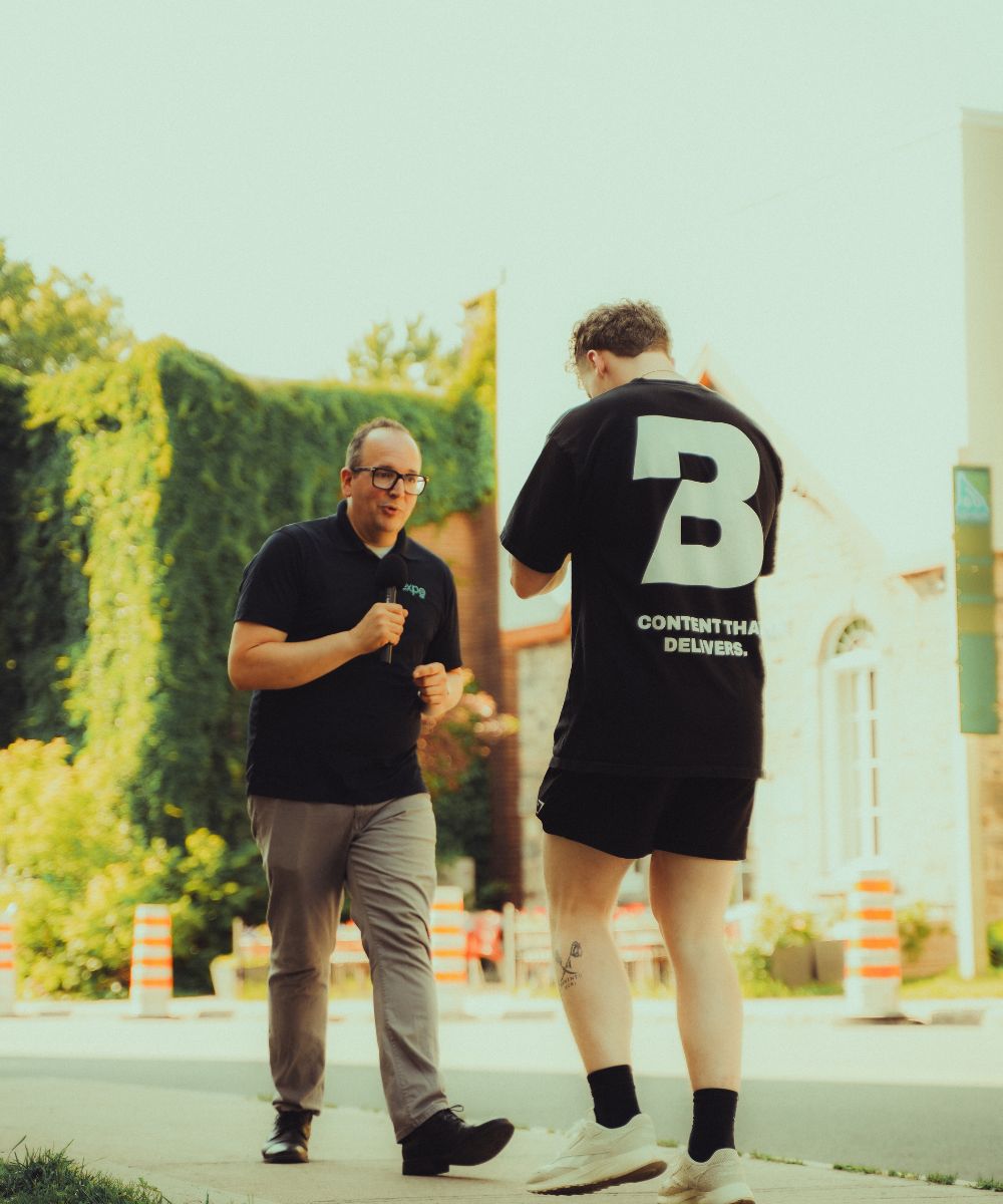 Two men standing on a sidewalk outdoors, one holding a microphone and the other wearing a black shirt with large white B and text on the back.