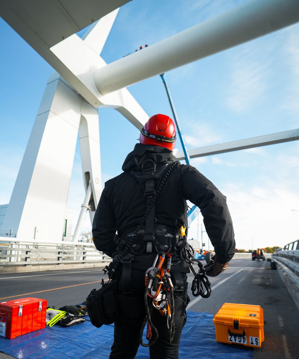 Worker wearing a red safety helmet and black gear stands on a bridge with climbing equipment attached, looking up at a large white structure and a crane.