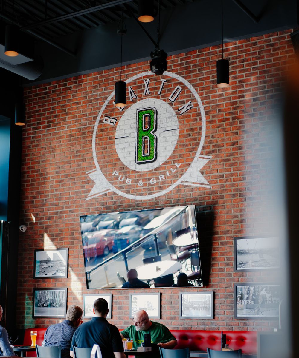 Interior of Braxton Pub & Grill with brick wall, large TV, framed photos, and people sitting at a table.