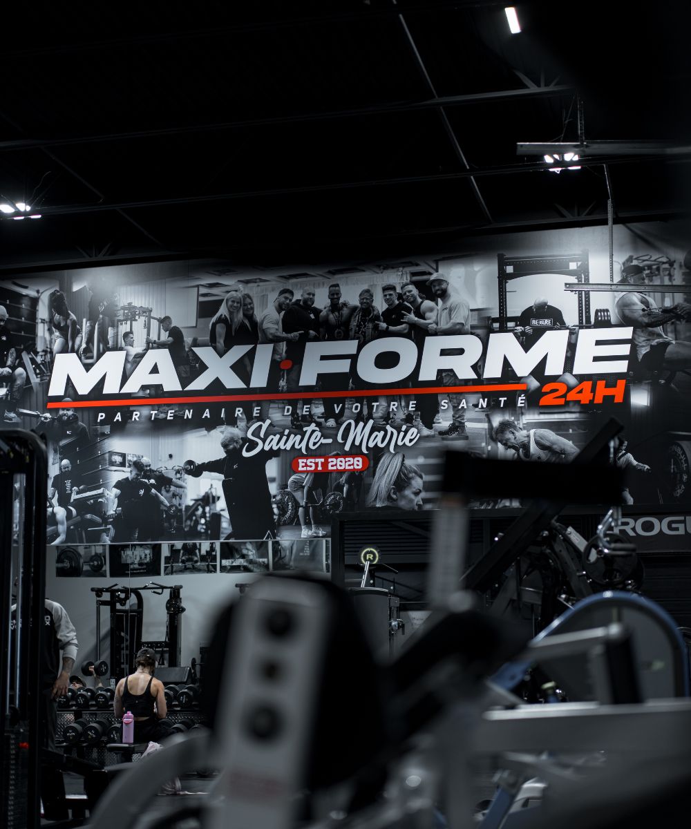 Gym interior with weightlifting equipment and a large wall mural featuring people exercising and the text 'MAXI FORME 24H Partenaire de Votre Santé Sainte-Marie EST 2020'.