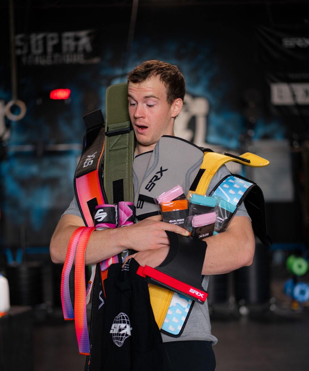 Young man in a gym holding multiple colorful fitness straps and resistance bands, looking surprised.