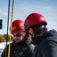 Two workers wearing red safety helmets and black jackets with a rope between them under a blue sky.