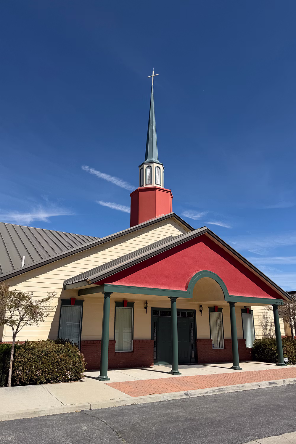 Small church building with beige walls, red brick base, green columns, and a tall steeple topped with a cross under a clear blue sky.