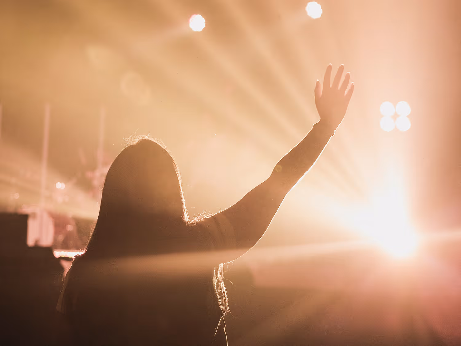 Person raising hand in a warmly lit concert or worship setting with bright stage lights shining in the background.
