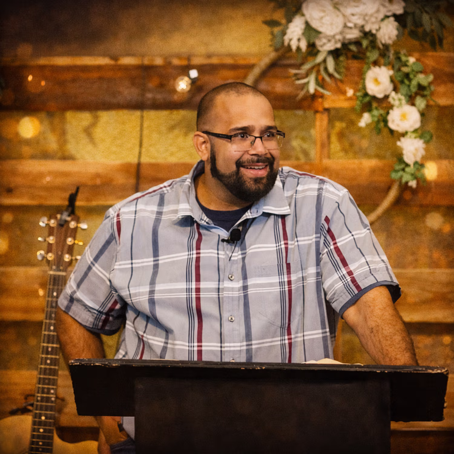 Pastor Indy speaking at a podium with a guitar and floral decoration in the background.