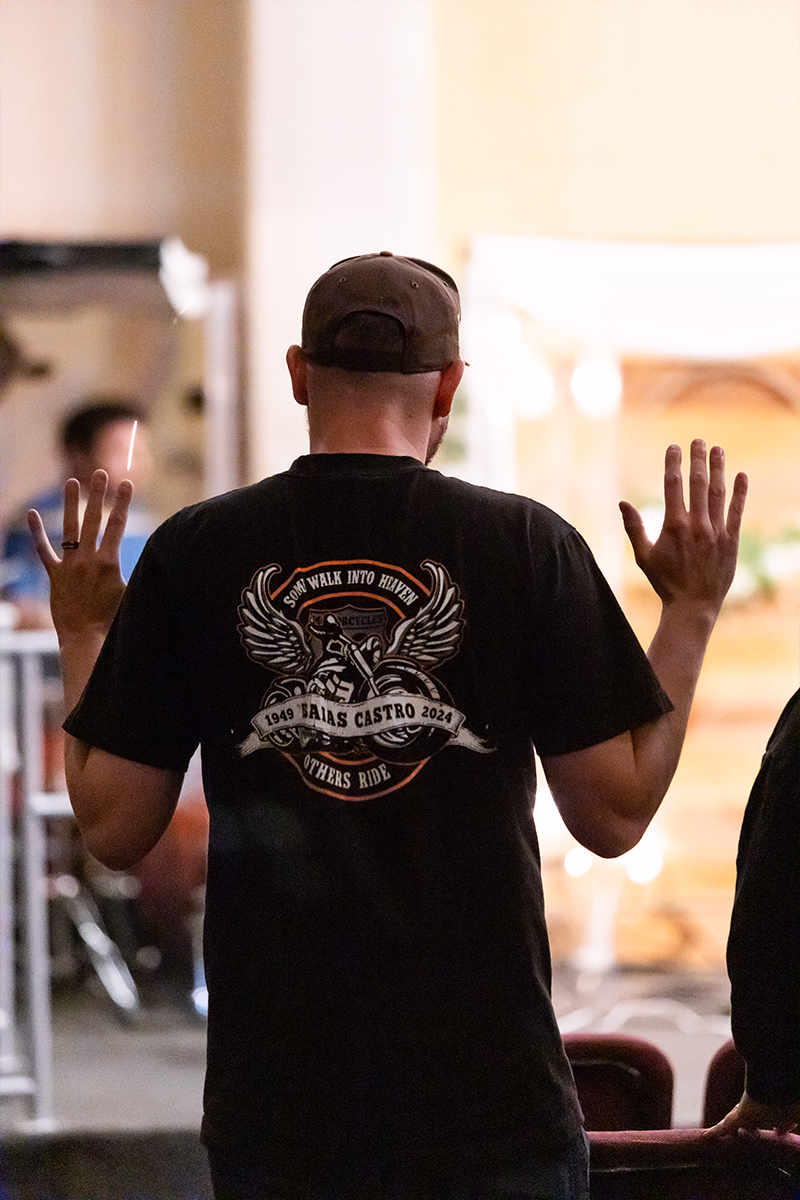 Man facing away wearing a black cap and T-shirt with motorcycle memorial design, hands raised.