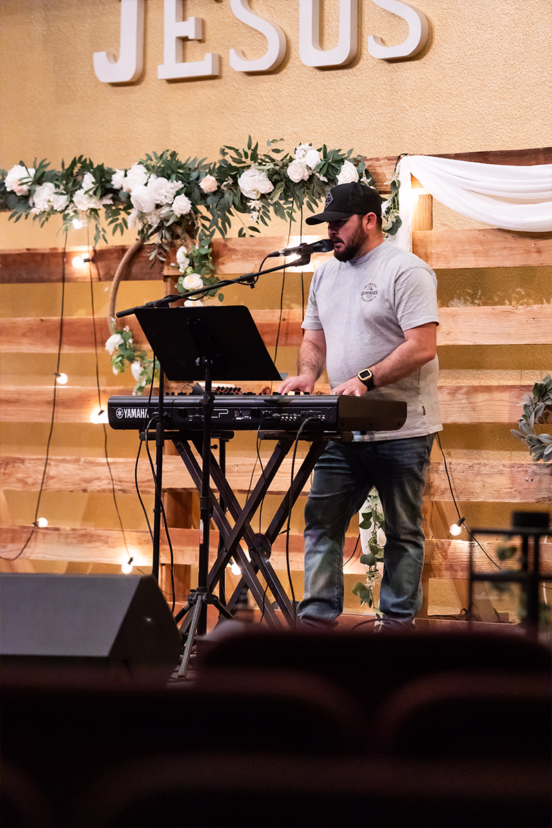 Man wearing cap playing a Yamaha keyboard and singing on stage decorated with flowers and lights under large JESUS letters.