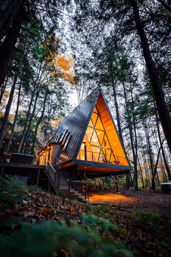 A-frame cabin with large glass windows glowing warmly, set on stilts in a forest during autumn.