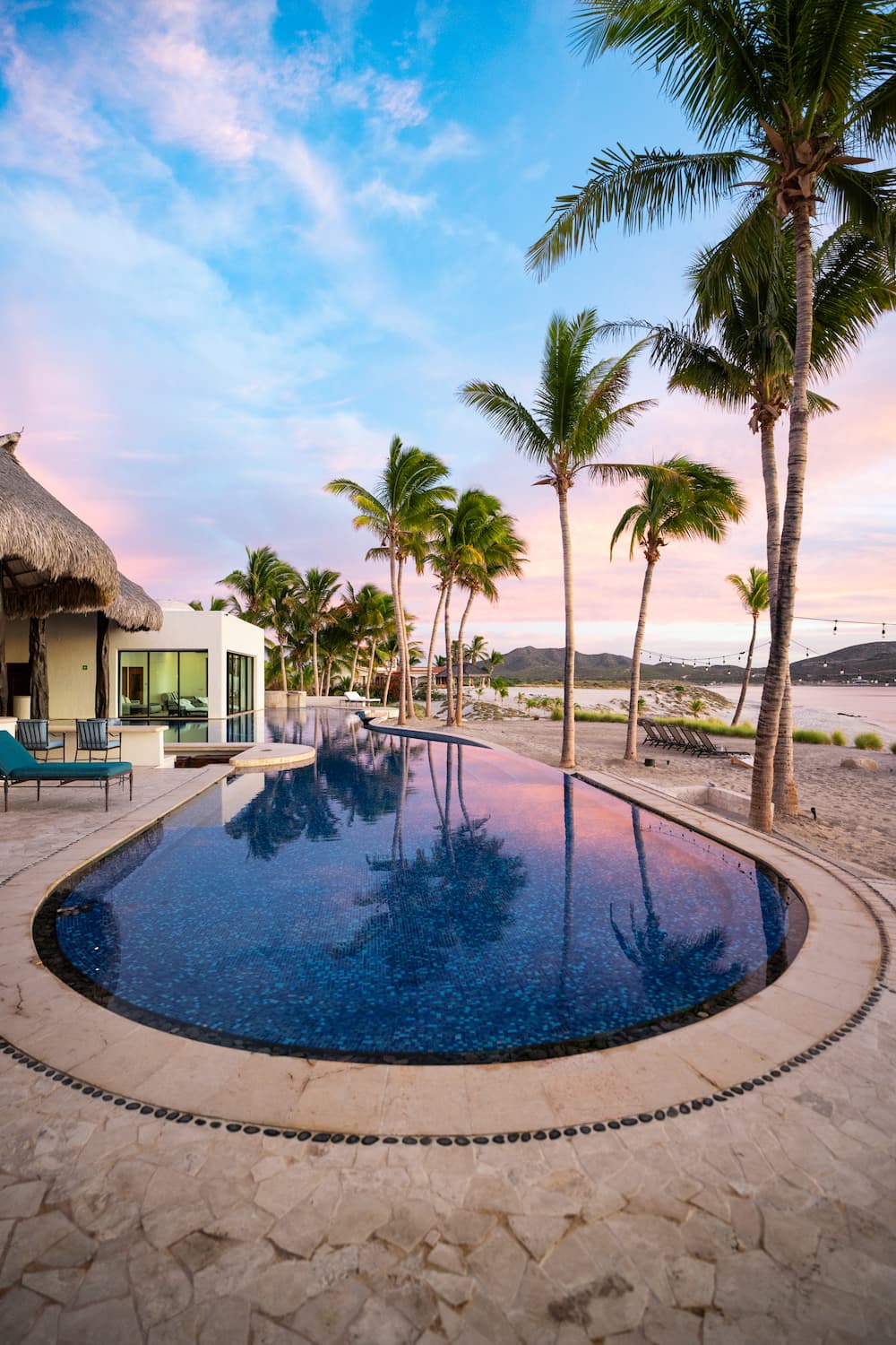 Infinity pool beside beach with palm trees, lounge chairs, and a colorful sunset sky.