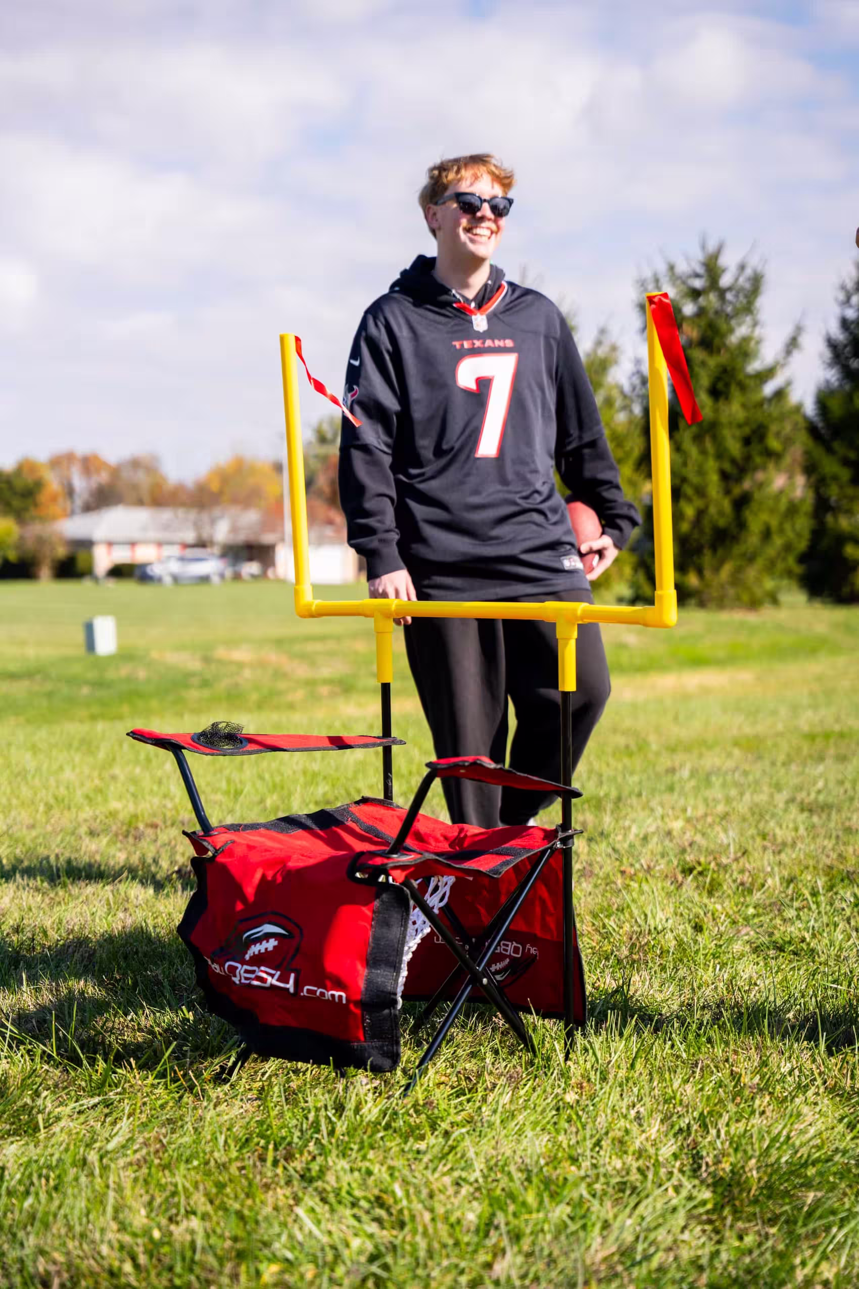 Young person wearing Texans jersey holding football behind yellow mini goalpost on green field.