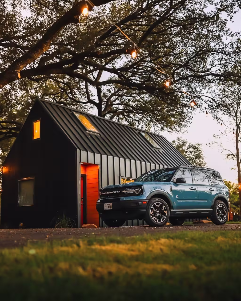 Blue SUV parked in front of a modern black house with warm lights under large trees at dusk.