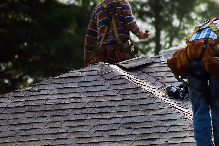 Two roofers wearing tool belts working together to lay overlapping asphalt shingles on steep residential rooftop.