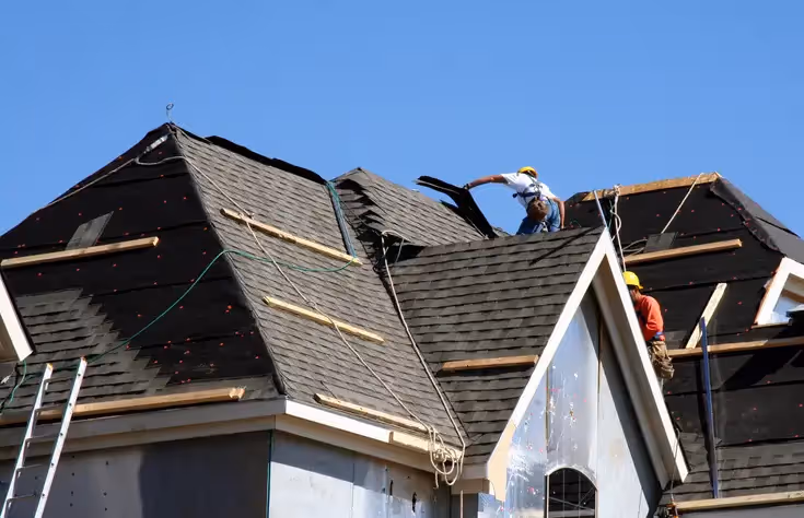 Construction worker in yellow helmet removing old black shingles from a steep residential roof under clear blue sky.