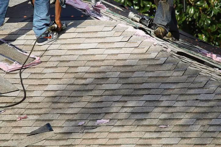 Worker using nail gun to install light gray asphalt shingles on roof, pink underlayment visible underneath.