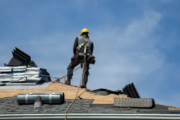 Roof worker in yellow hard hat and safety harness standing on sloped roof with materials stacked nearby.