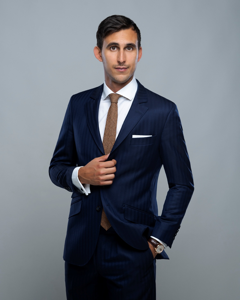 Homme portant un costume bleu marine rayé avec une cravate marron et une montre-bracelet, posant devant un fond gris.