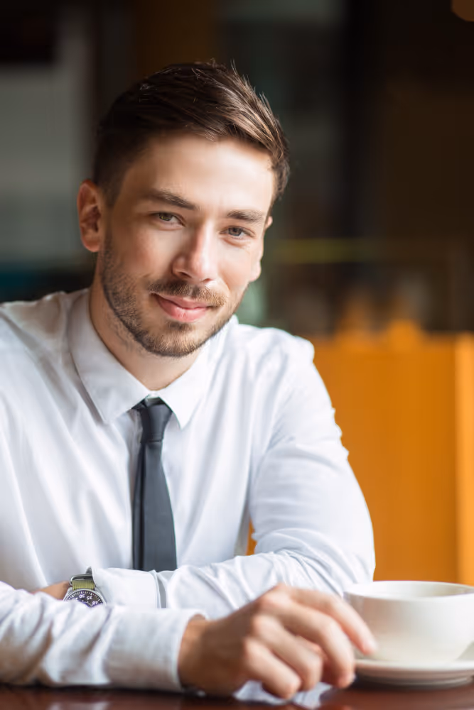 Young man in a white shirt and black tie sitting at a table with a cup of coffee.