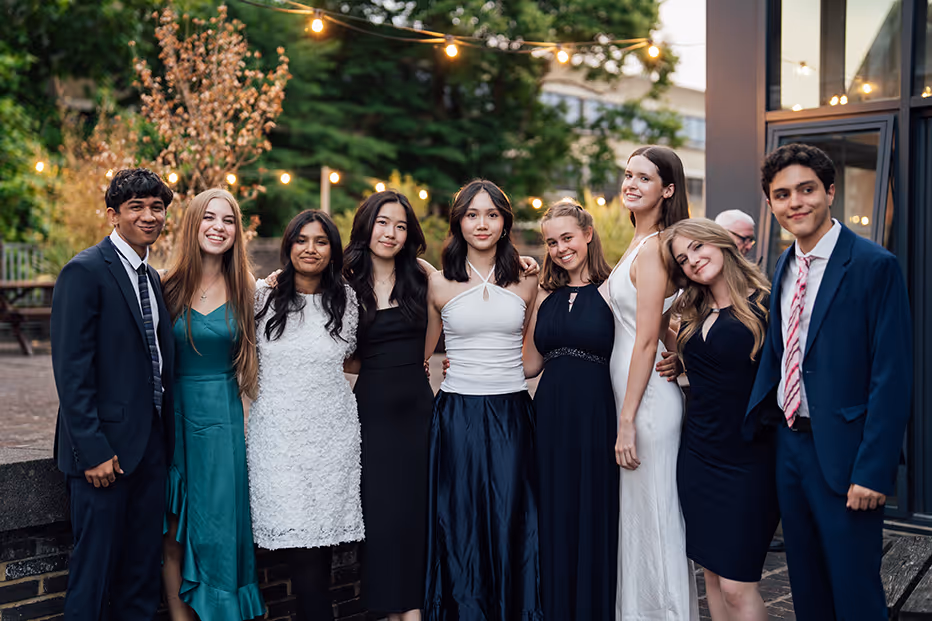 Group of eight young adults dressed formally, posing outdoors with string lights and greenery in the background.
