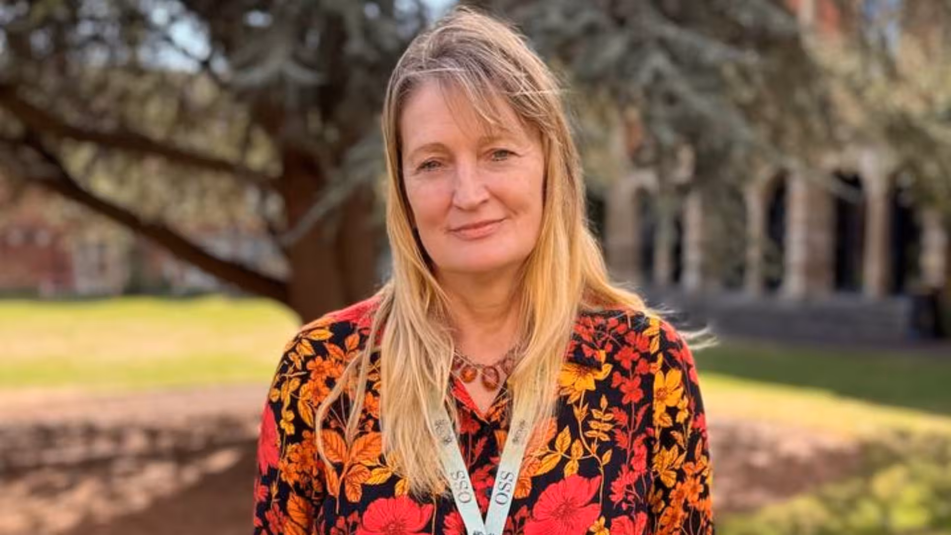 Woman with long blonde hair wearing a floral top and lanyard standing outdoors with trees and grass in the background.