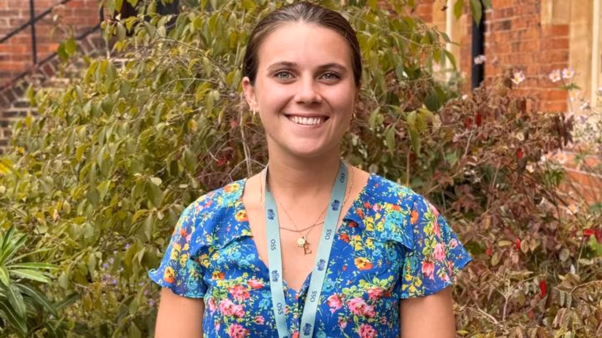 Smiling woman wearing a blue floral dress and light blue lanyard standing in front of leafy plants and a brick building.