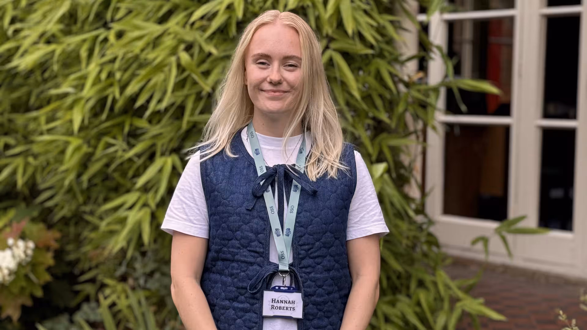 Smiling young woman with blonde hair wearing a white shirt and blue quilted vest, standing outdoors with greenery and a windowed door behind her.