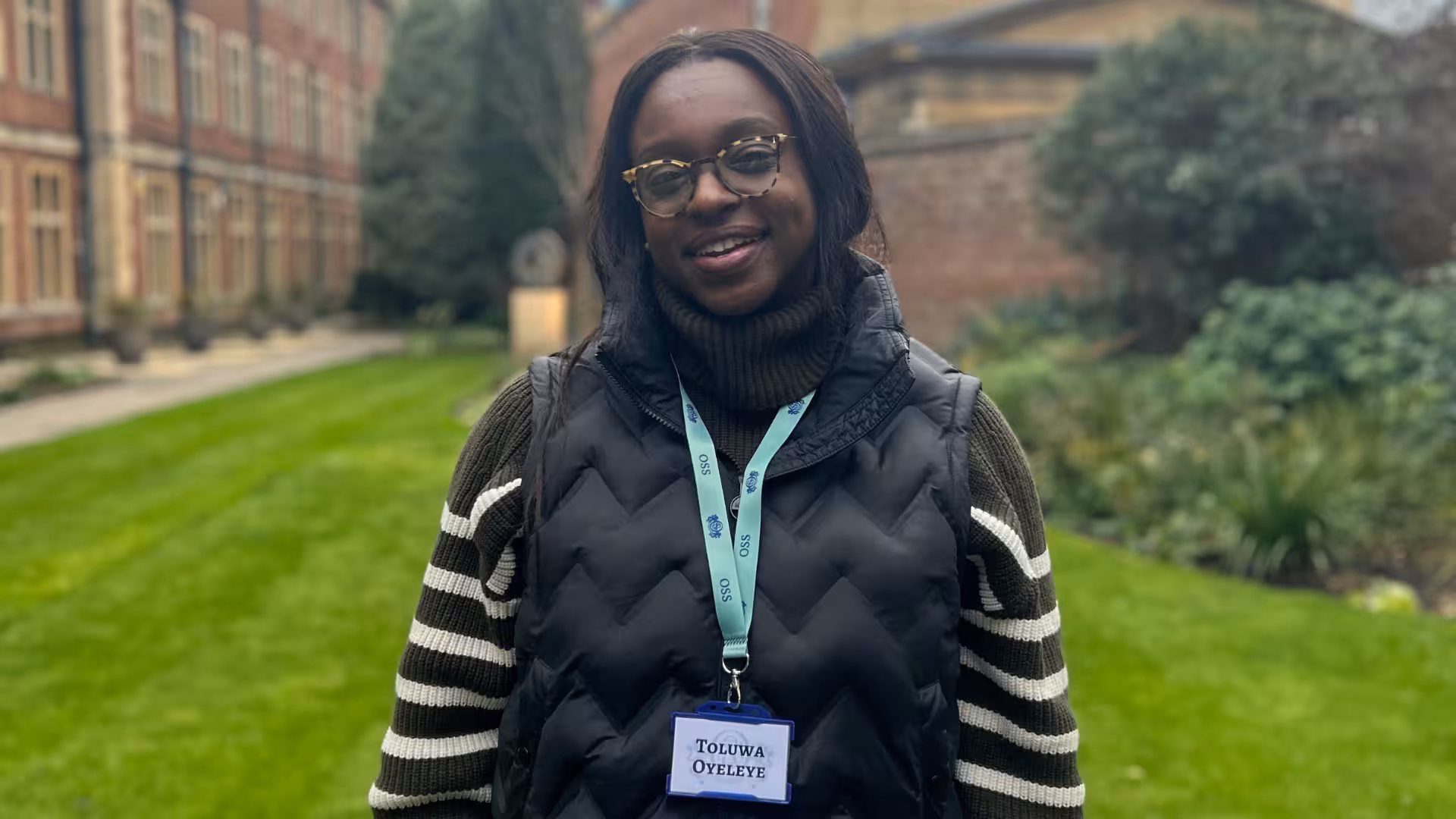 Smiling woman wearing glasses, a black puffer vest, and striped sweater standing outdoors with green grass and buildings in the background.