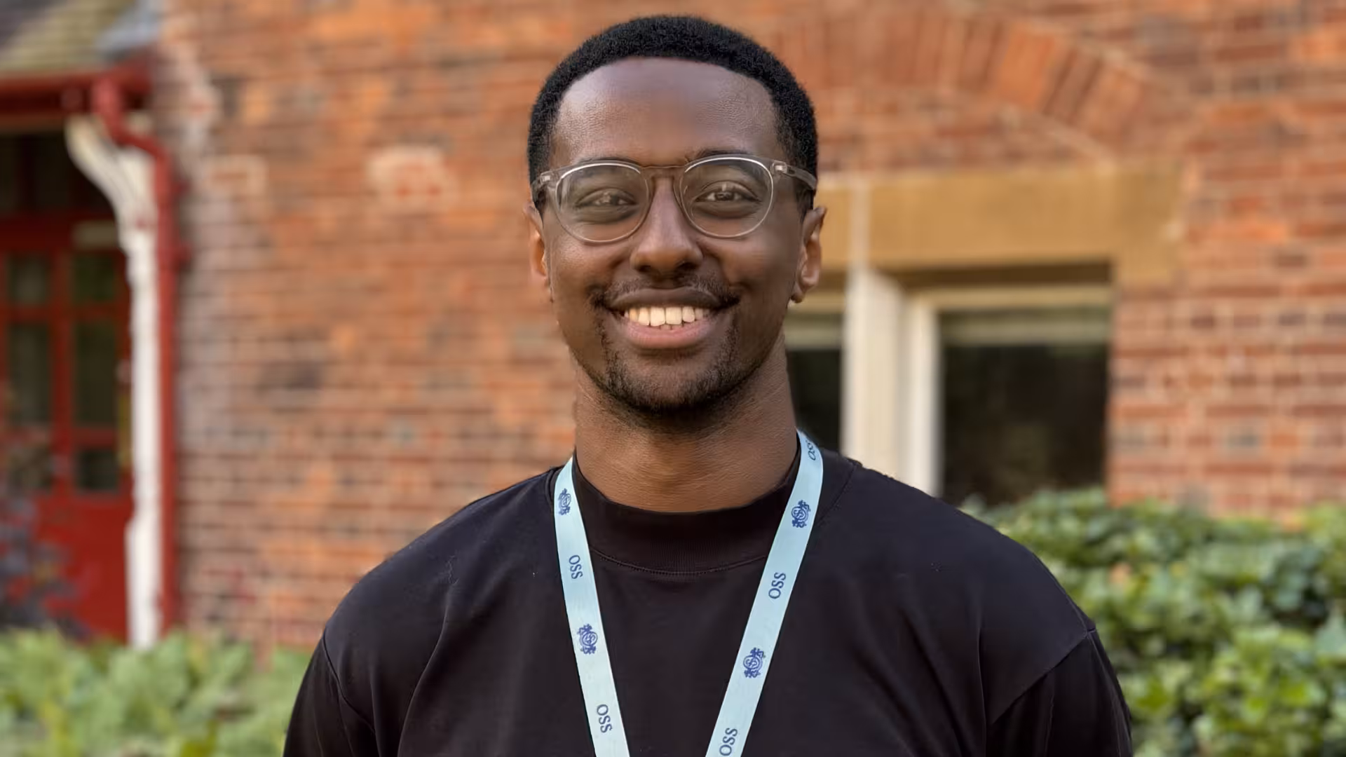 Smiling man wearing glasses and a light blue lanyard standing outdoors in front of a brick building.