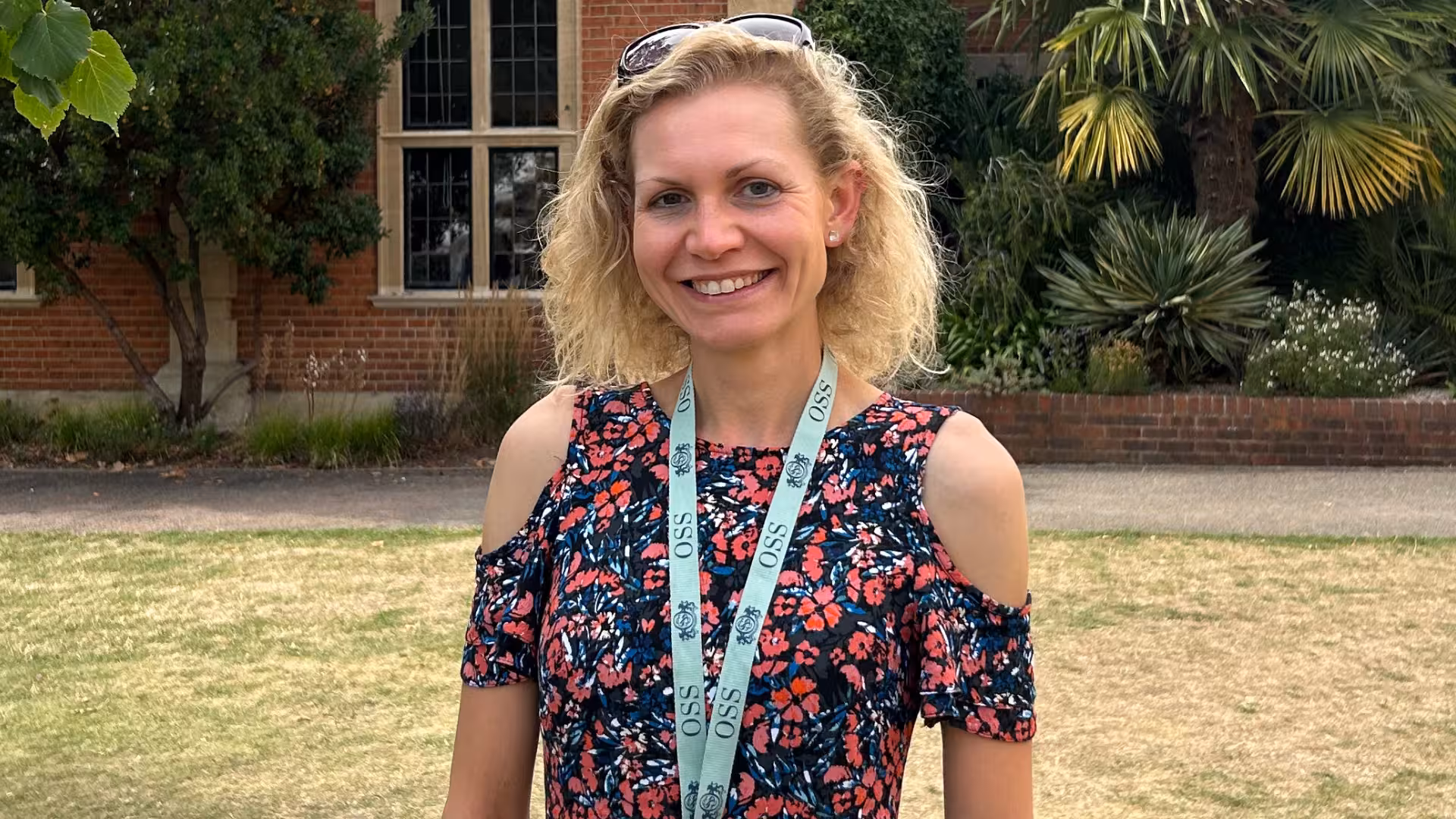 Smiling woman with curly blonde hair wearing sunglasses on her head and a floral cold-shoulder top outdoors in a garden.