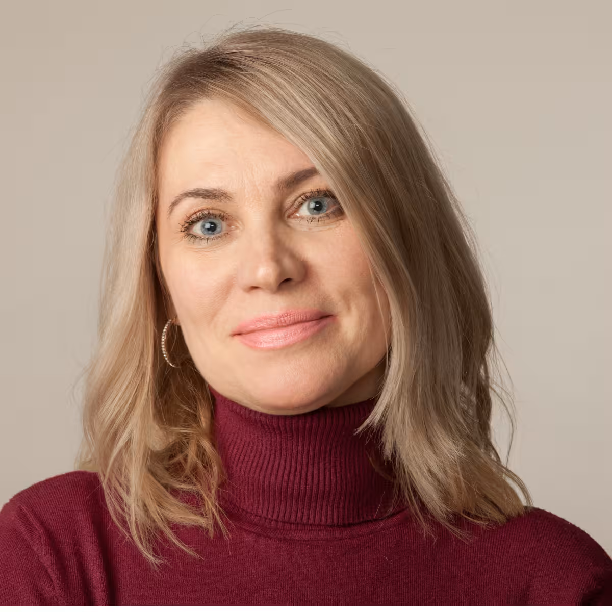 Smiling woman with blonde hair wearing a burgundy turtleneck sweater and hoop earrings against a beige background.