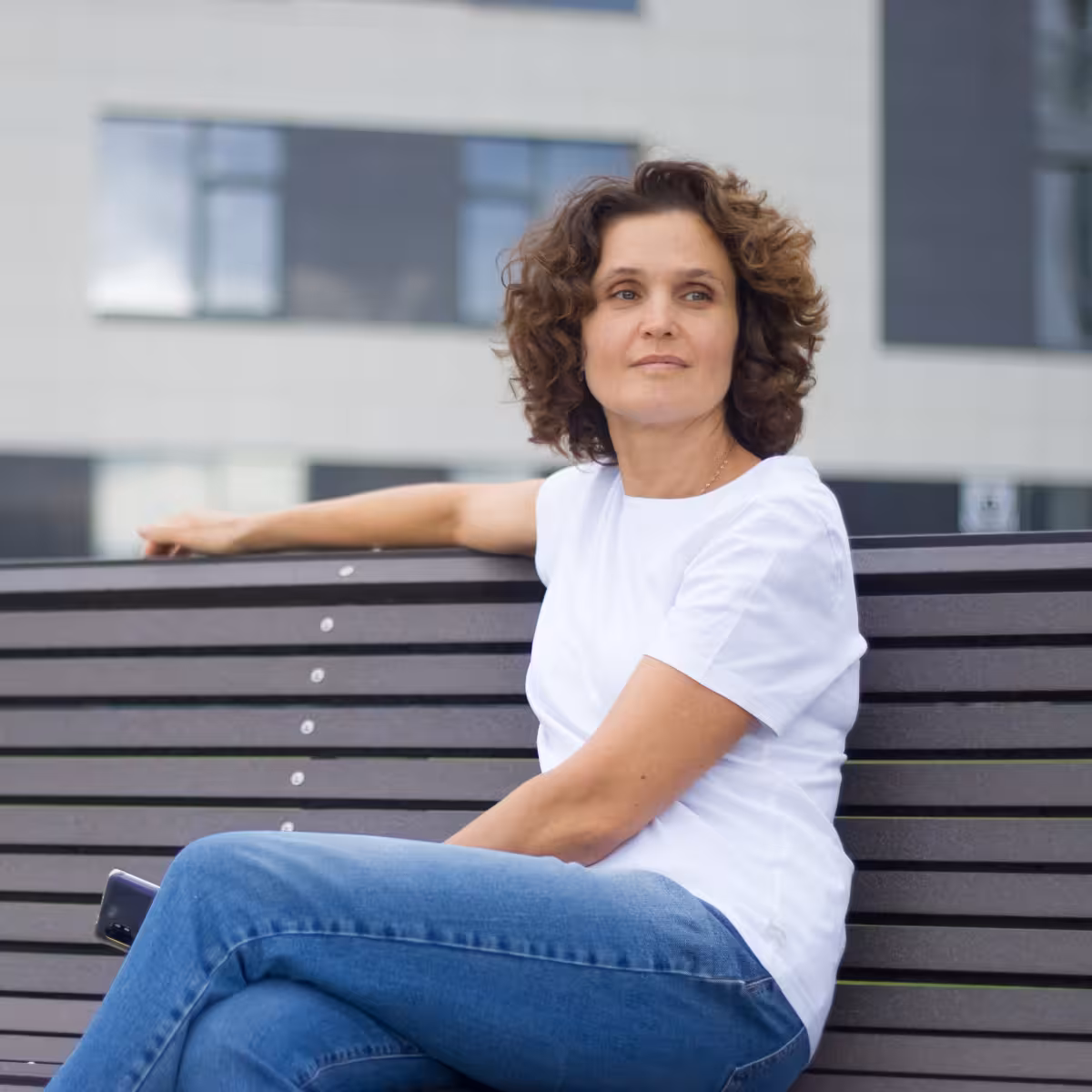 Woman with curly hair wearing a white t-shirt and blue jeans sitting on a bench outdoors.