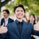 Smiling young man in a suit giving a thumbs-up outdoors with blurred friends in the background.